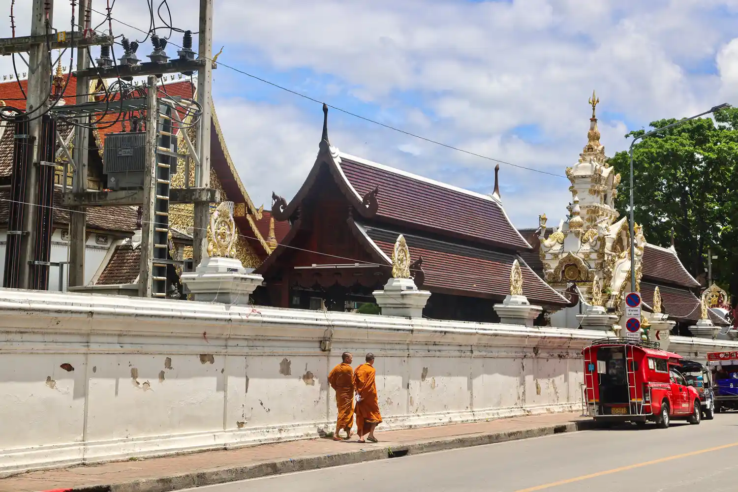 Two monks in orange robes walking past a traditional Thai wall and temple architecture in Chiang Mai.