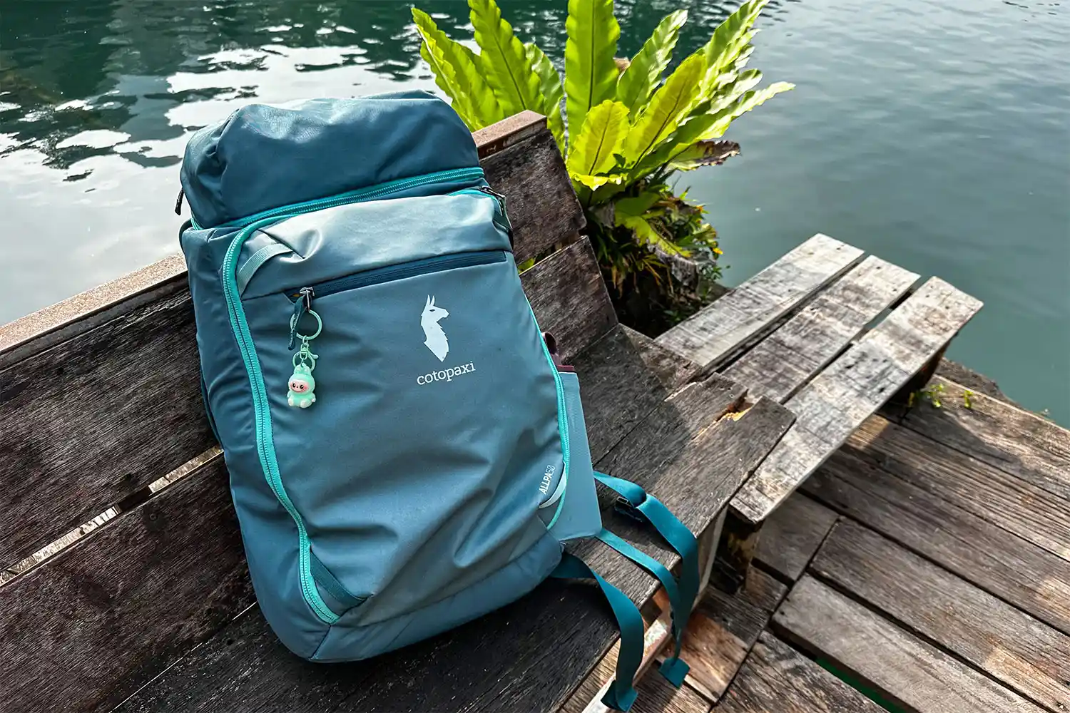 A Cotopaxi travel backpack resting on a wooden bench near a body of water.
