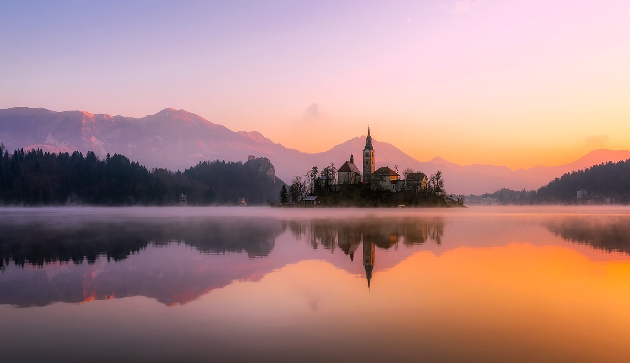 A modern A-frame cabin reflecting in a clear mountain lake with snow-capped peaks in the background.