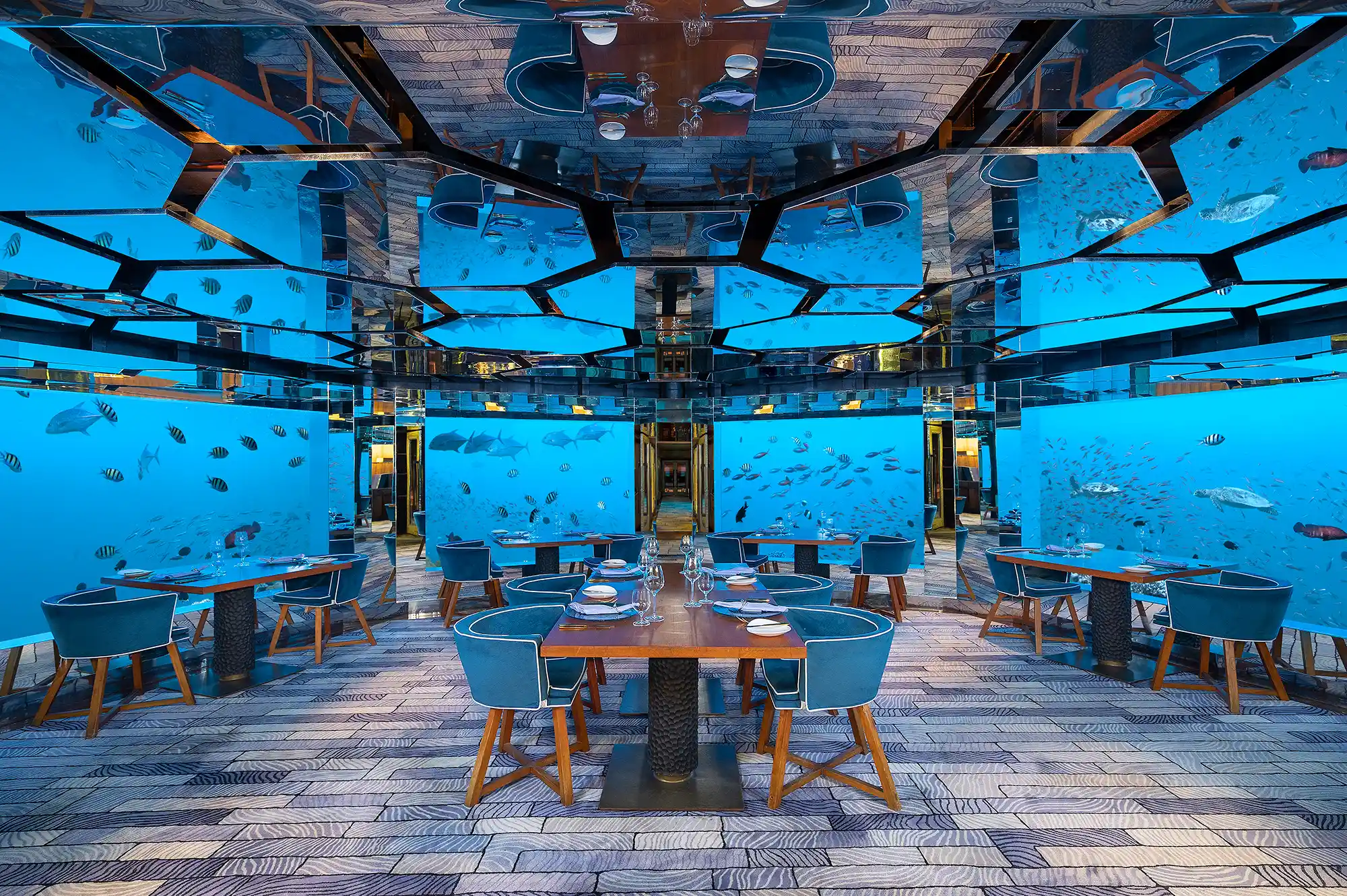 Fine dining tables set inside an underwater restaurant surrounded by marine life and coral.