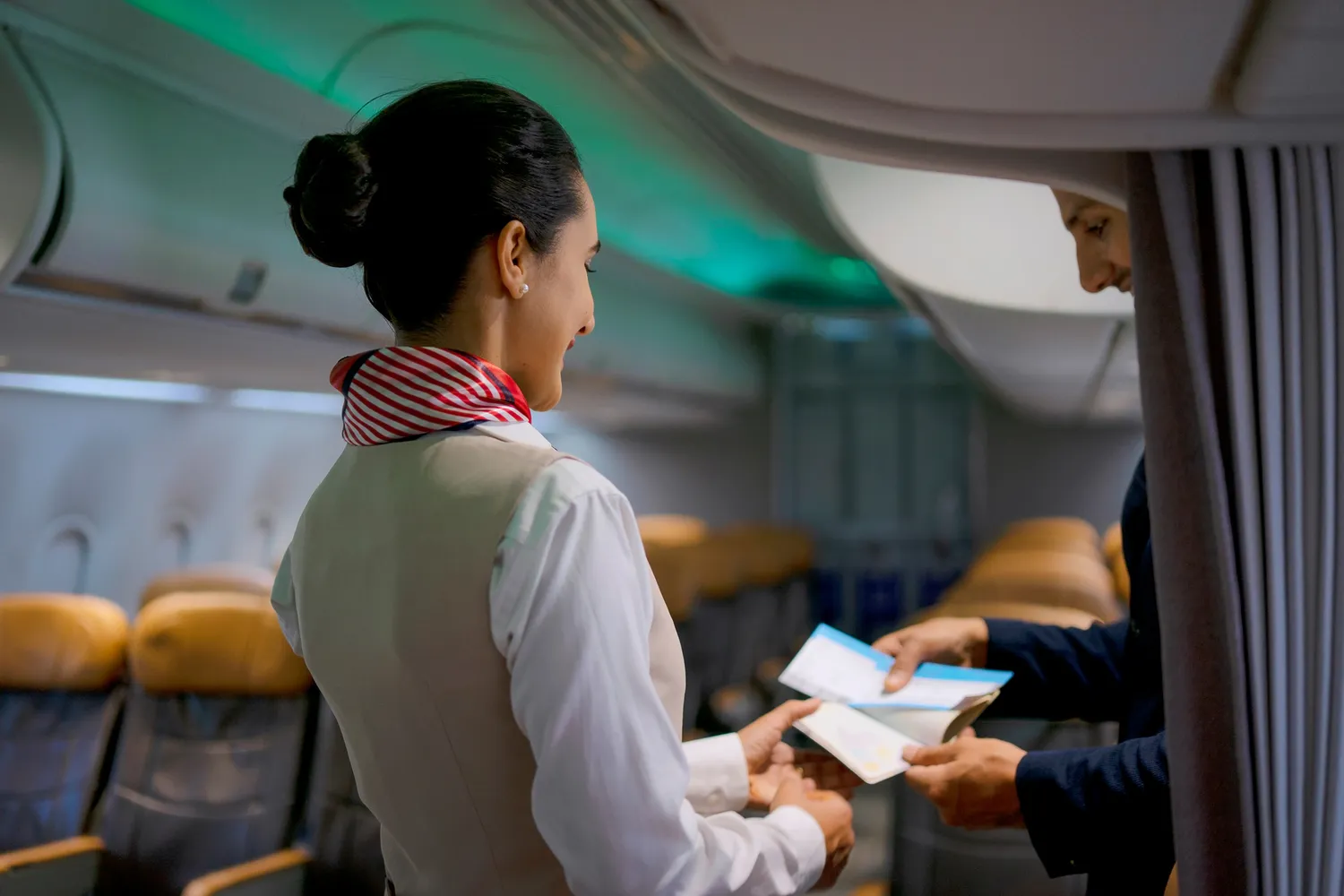 A flight attendant's hands receiving a boarding pass from a passenger in the airplane cabin.