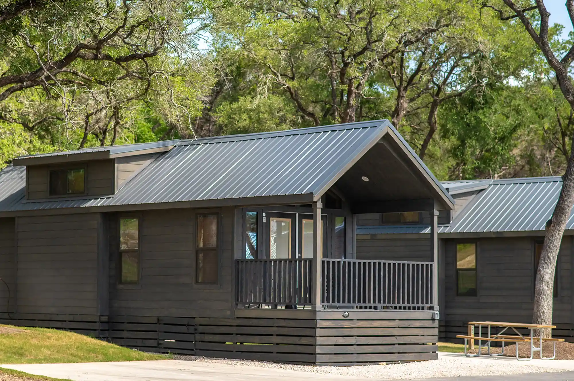 A modern wooden cabin with a porch nestled among shaded trees at a campground.