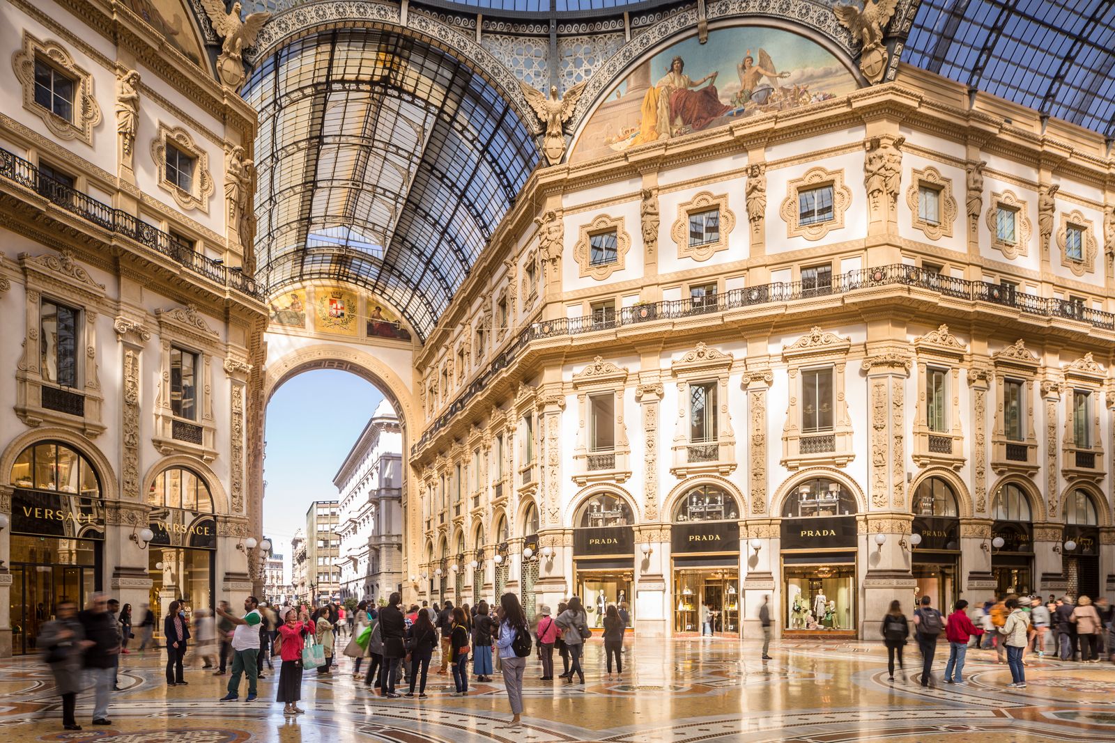 Shoppers walking through a grand, glass-vaulted historic shopping arcade in Milan.
