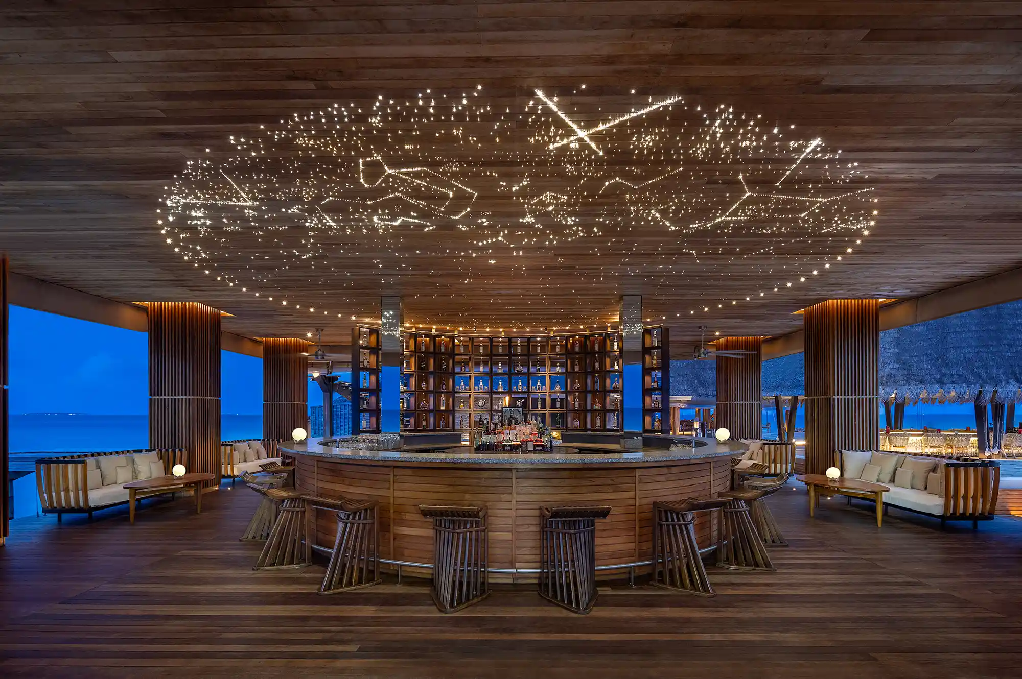 A modern circular bar under a ceiling featuring constellations, looking out over the evening ocean.