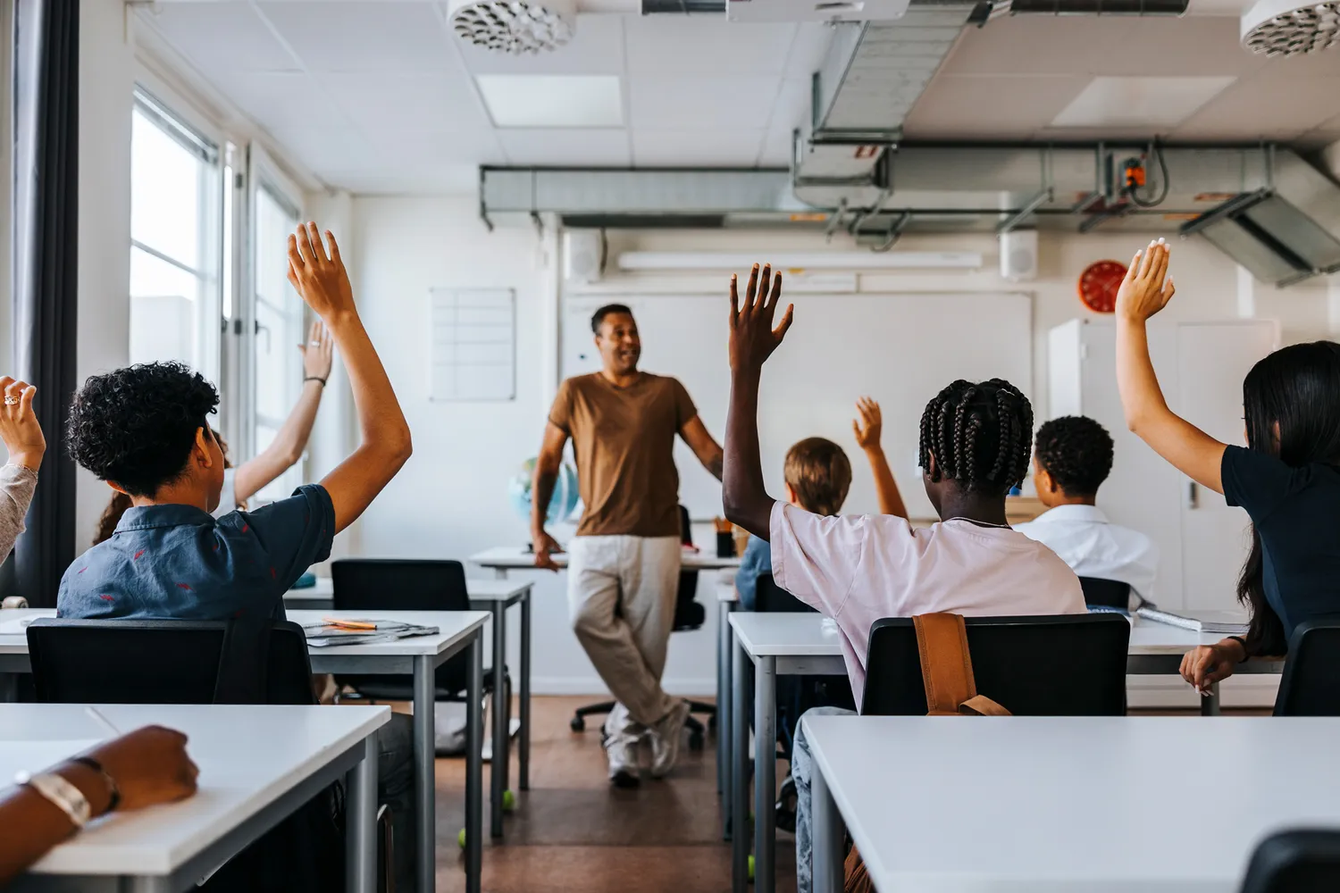 A teacher standing in front of a classroom with students raising their hands.