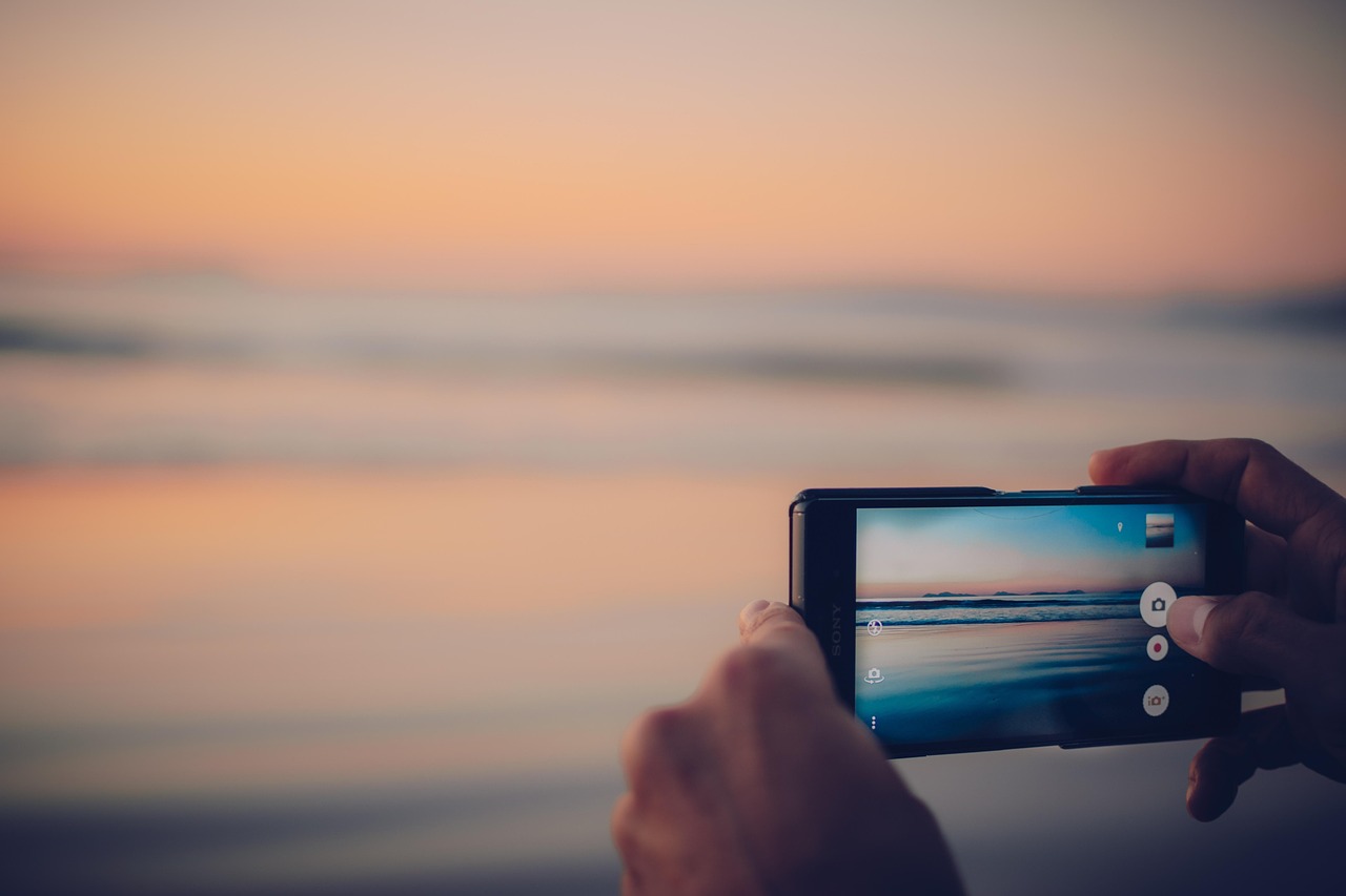 A person holding a smartphone with an external wide-angle lens attached, overlooking a mountain range.