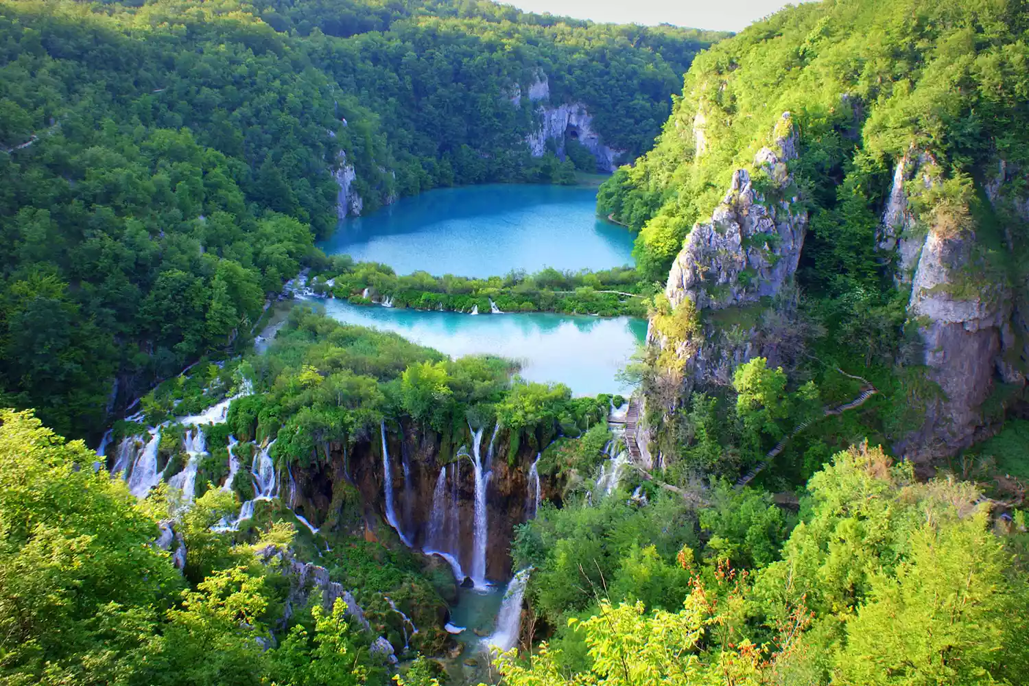 Multiple waterfalls flowing into crystal clear emerald lakes surrounded by dense forest in Plitvice, Croatia.