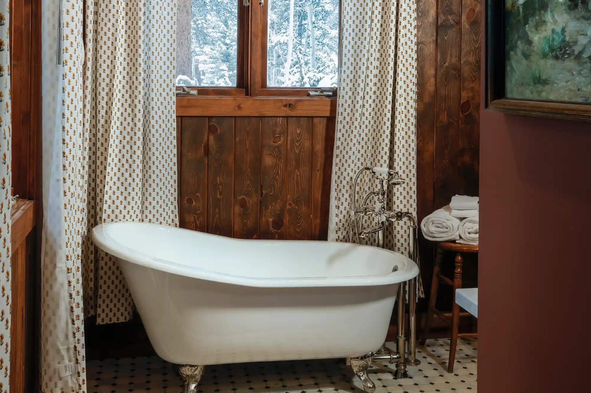 A rustic bathroom interior with a vintage-style freestanding claw-foot bathtub set against warm wooden plank walls and a forest-view window.