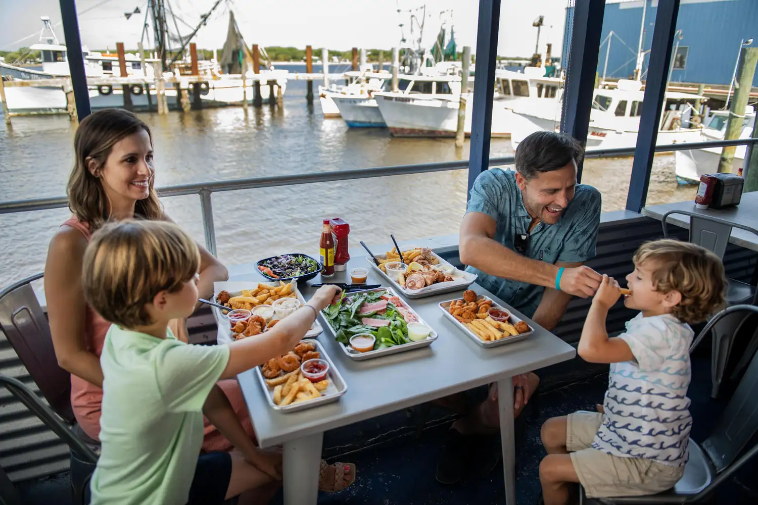 A family dining outdoors at a seafood market with fishing boats visible in the background.
