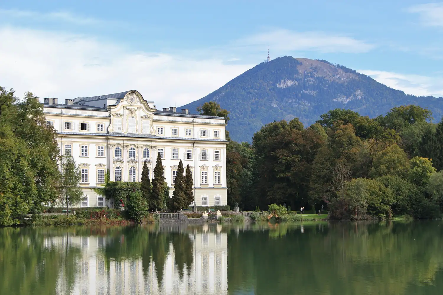 Leopoldskron Palace reflected in the calm waters of the lake with mountains in the distance.