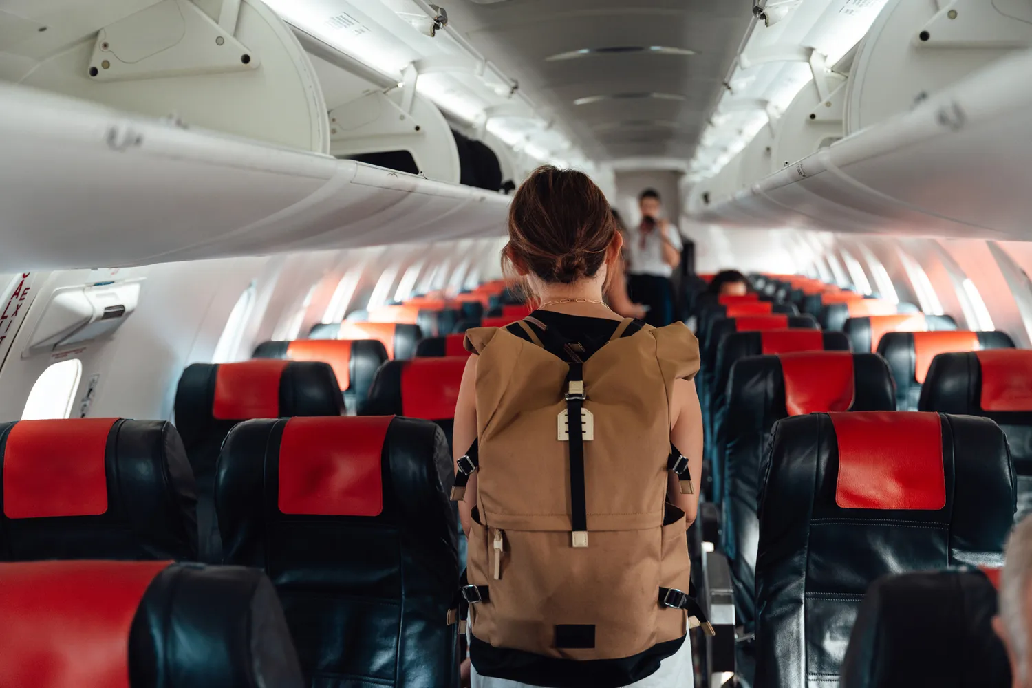 A young woman carrying a backpack walking down an empty airplane aisle with red and black seats.