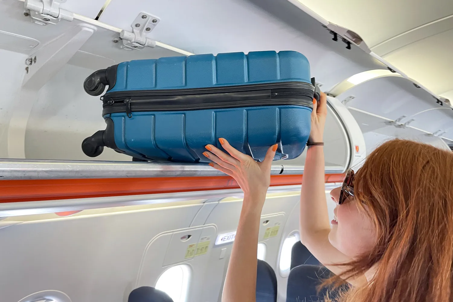 A passenger lifting a black roller bag into an airplane's overhead bin with wheels facing inward.