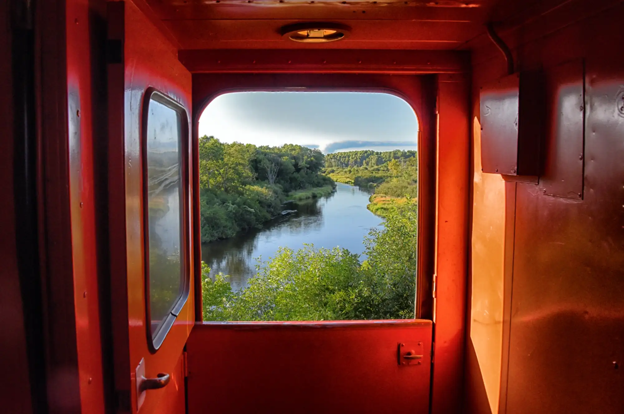 View of a serene river and landscape seen through the open rear doorway of a train car.