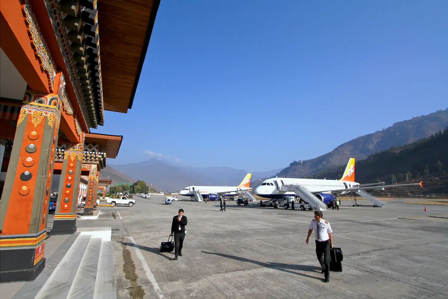 Two airline crew members walking on the tarmac at Paro International Airport in Bhutan with mountains in the distance.