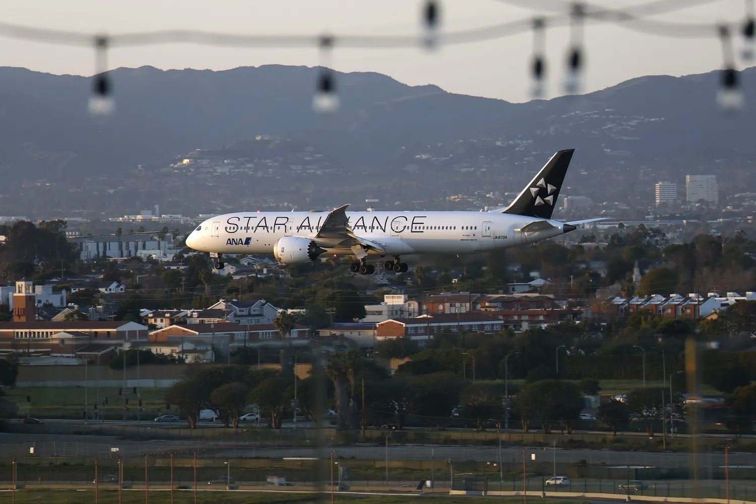 An All Nippon Airways airplane descending toward a runway with a mountainous city background.