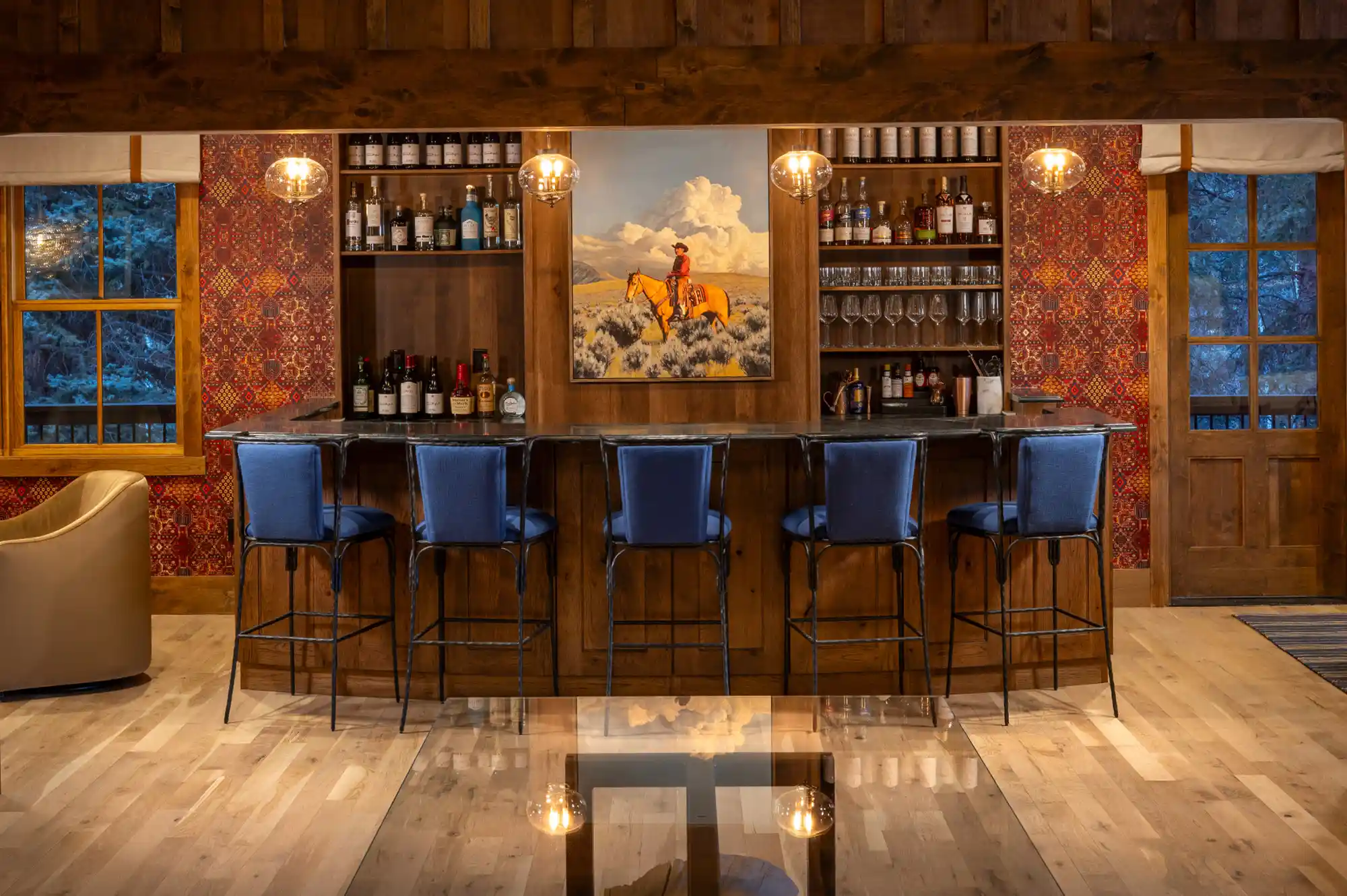 A stylish lodge bar area with blue velvet stools and a prominent oil painting of a cowboy on horseback.