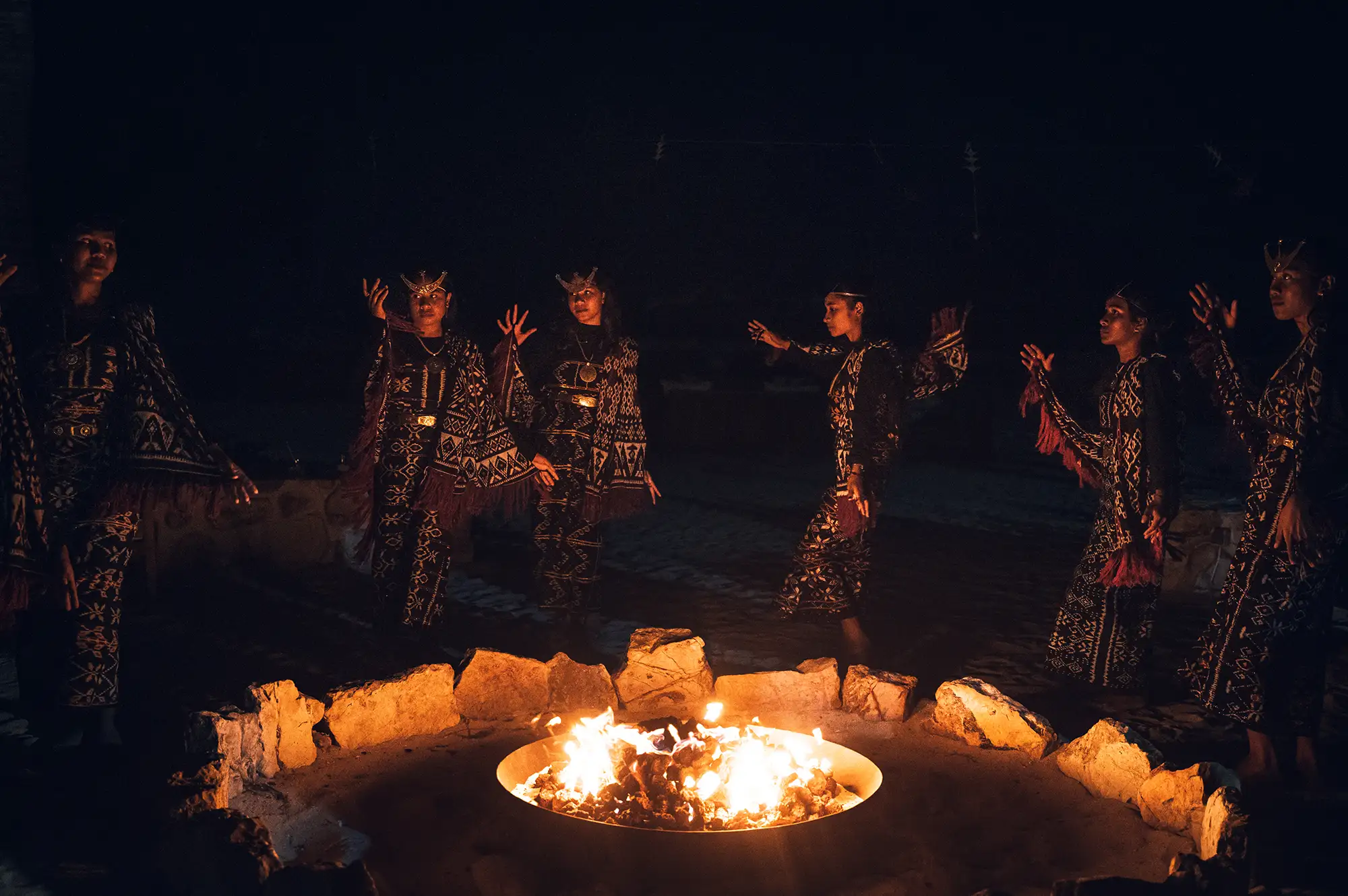 Performers in traditional Rotenese ikat costumes dancing around a fire pit at night.