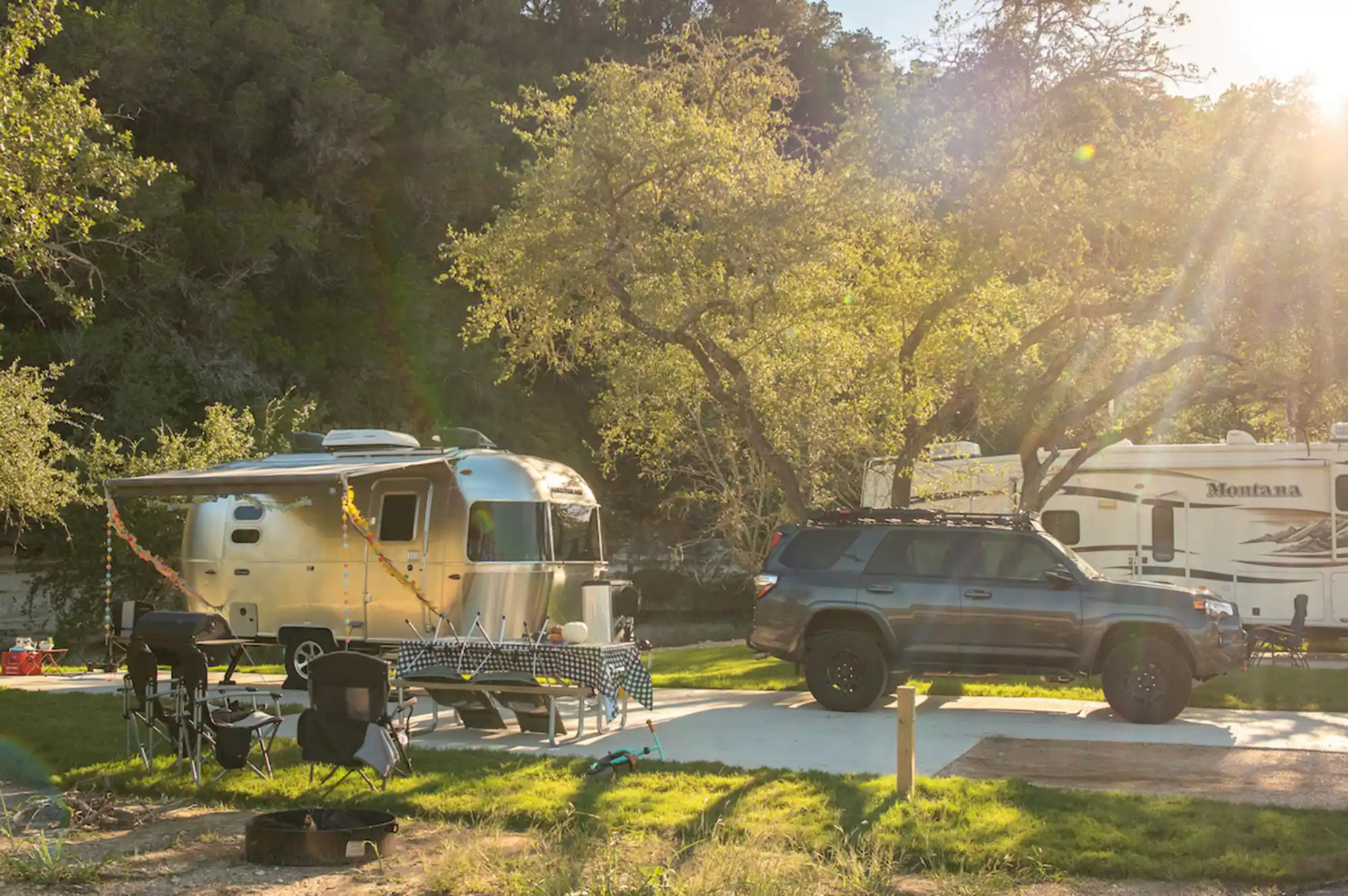 A silver Airstream-style trailer parked at a wooded campsite with outdoor seating in the sun.